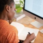 woman writing in her notebook sitting in front of a computer monitor