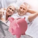 smiling retirees relaxing in bed with a pink piggy bank in the foreground representing their nest egg